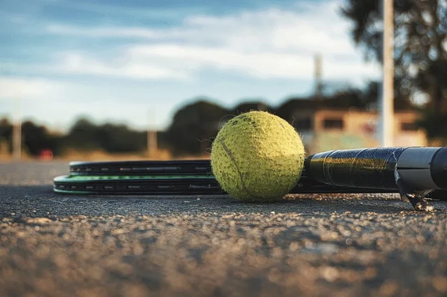 Tennis ball and rackets resting on an outdoor court, surrounded by blurred trees in the background, evoke a sense of performance impact common among sports athletes.
