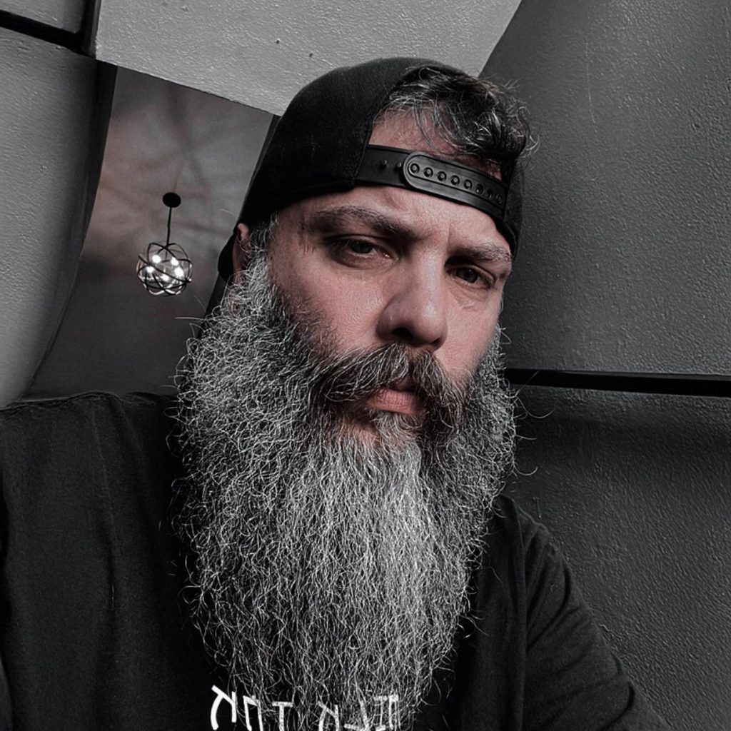 Man with a long gray beard and backward black cap, wearing a black shirt, sits in a dimly lit room.