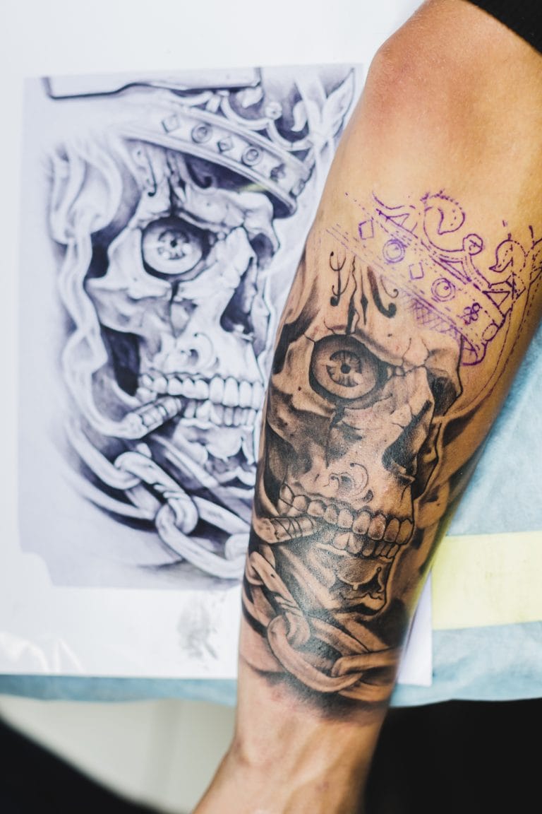 Forearm tattoo in progress by Funhouse Artists features a crowned skull masterpiece; a stencil and reference sketches of the same skull are placed beside the arm.