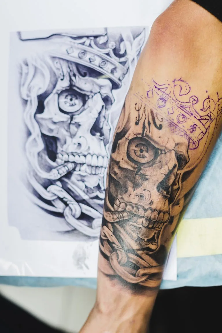 Forearm tattoo in progress by Funhouse Artists features a crowned skull masterpiece; a stencil and reference sketches of the same skull are placed beside the arm.