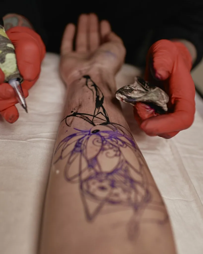 A person wearing red gloves begins tattooing a black ink design on someone's forearm, with the tattoo stencil visible—perfect for showcasing the benefits of plant-based tattoo aftercare or nourishing tattoo balms.
