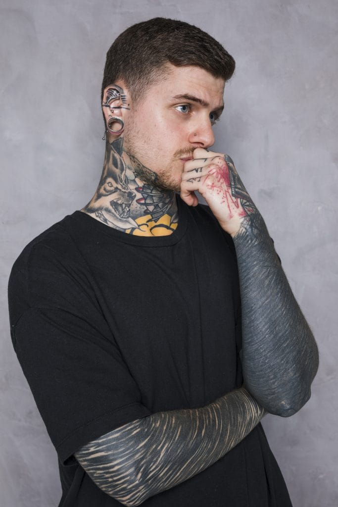 A man with tattoos on his neck, arms, and hand—some featuring blast-overs and old ink—wears a black shirt as he poses with his hand on his chin against a gray background.