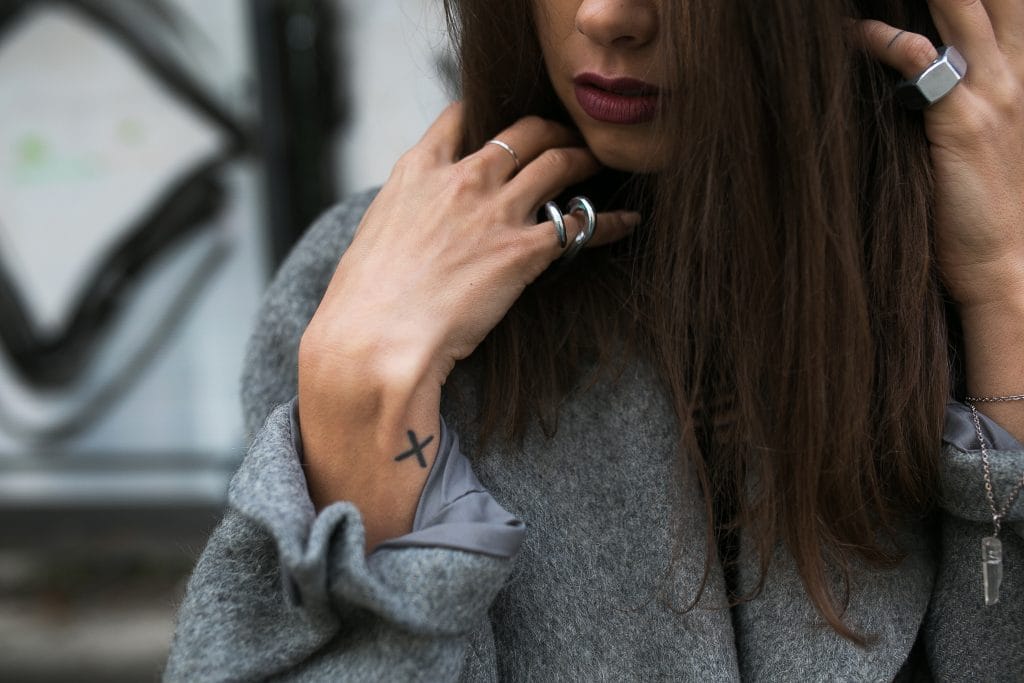 A woman with long brown hair wearing a gray coat and silver rings shows off a small "X" fine-line micro tattoo on her wrist.