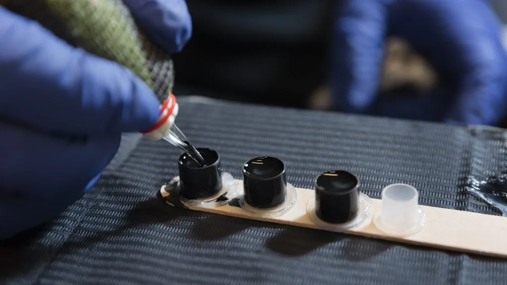 A gloved hand fills a small black ink cap with tattoo ink using a tattoo machine, next to other ink caps and a plastic container on a textured surface—key steps before considering tattoo placement and the possible pain scale.