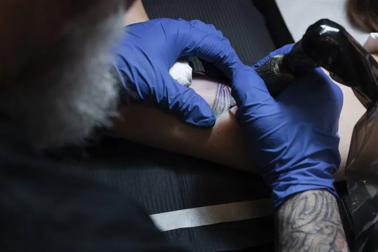 A tattoo artist wearing blue gloves works on a person's forearm with a tattoo machine, carefully considering tattoo placement and the potential tattoo pain for the client.