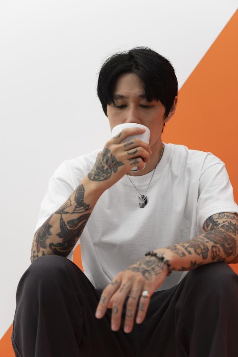 Person with tattoos sits against an orange and white background, wearing a white t-shirt, holding a cup near their mouth.