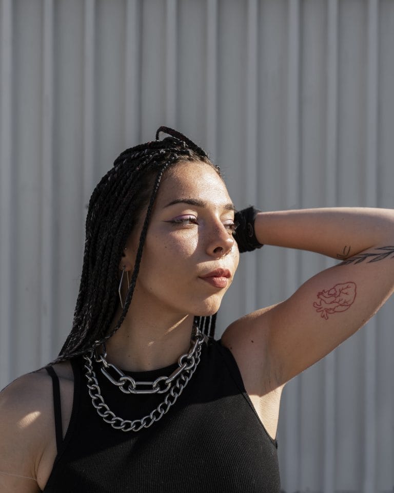 A woman with braided hair and a chain necklace poses outdoors, wearing a sleeveless black top and showing a tattoo on her upper arm.