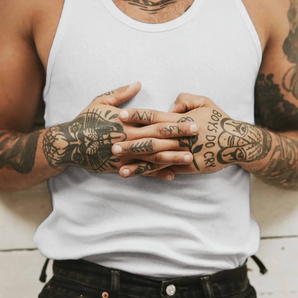 A person wearing a white tank top and black pants stands with tattooed hands clasped together; tattoos include a cat, a face, and the words "BOYS DO CRY.