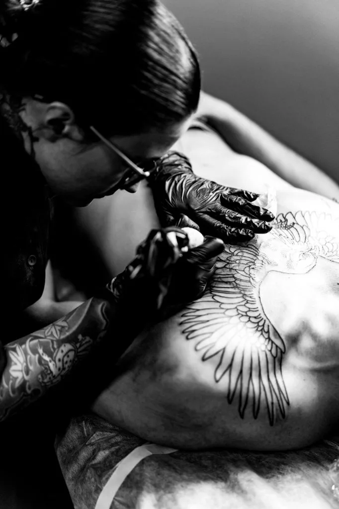A tattoo artist with gloves is tattooing a large winged design onto a person’s back in a black and white photo.