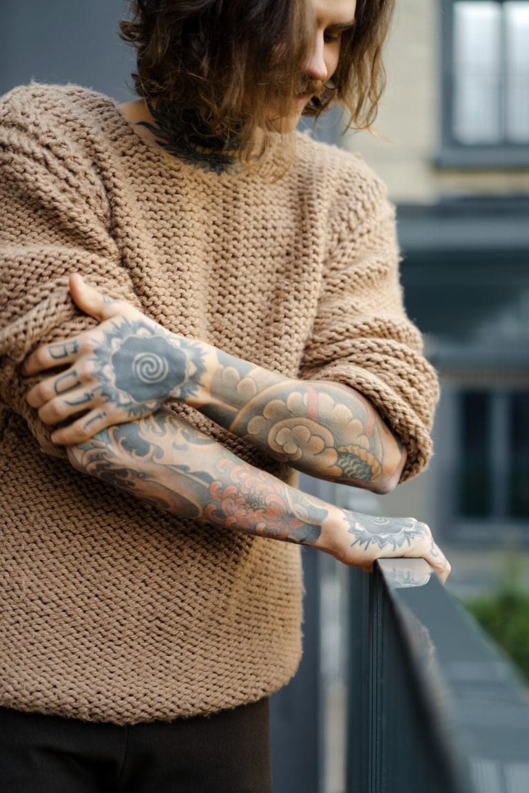 Person with long hair wearing a textured brown sweater, displaying tattooed arms while leaning on a railing outdoors.
