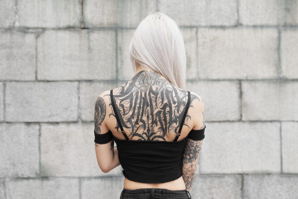A person with long, light blond hair stands facing a gray brick wall, showing extensive black tattoos covering their back, arms, and shoulders.