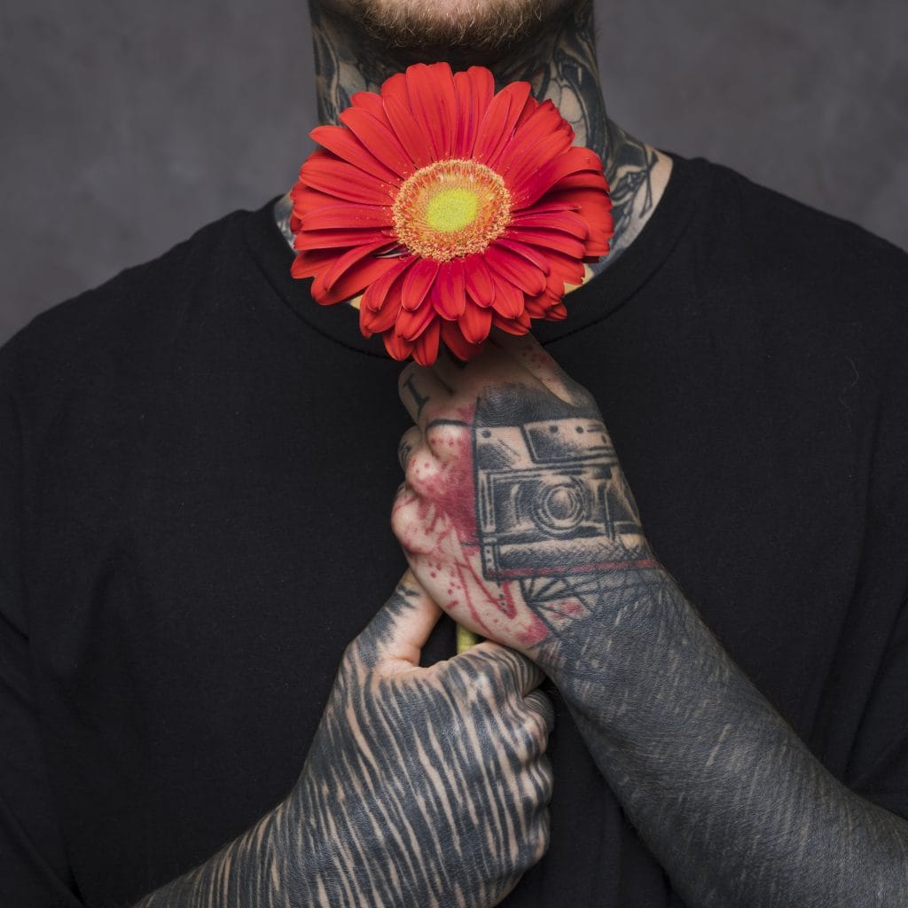 Person with tattooed arms and neck holds a red daisy flower to their chest, wearing a black shirt against a neutral background.
