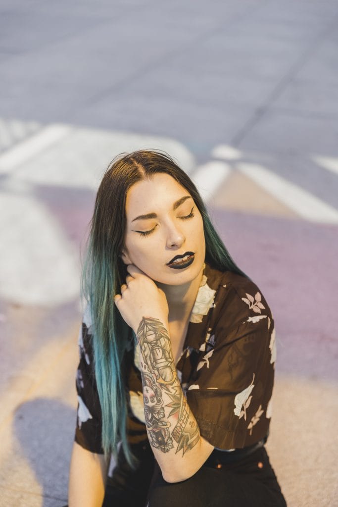 Woman with long teal hair, black lipstick, and a floral tattoo on her arm sits outdoors with eyes closed, resting her head on her hand.