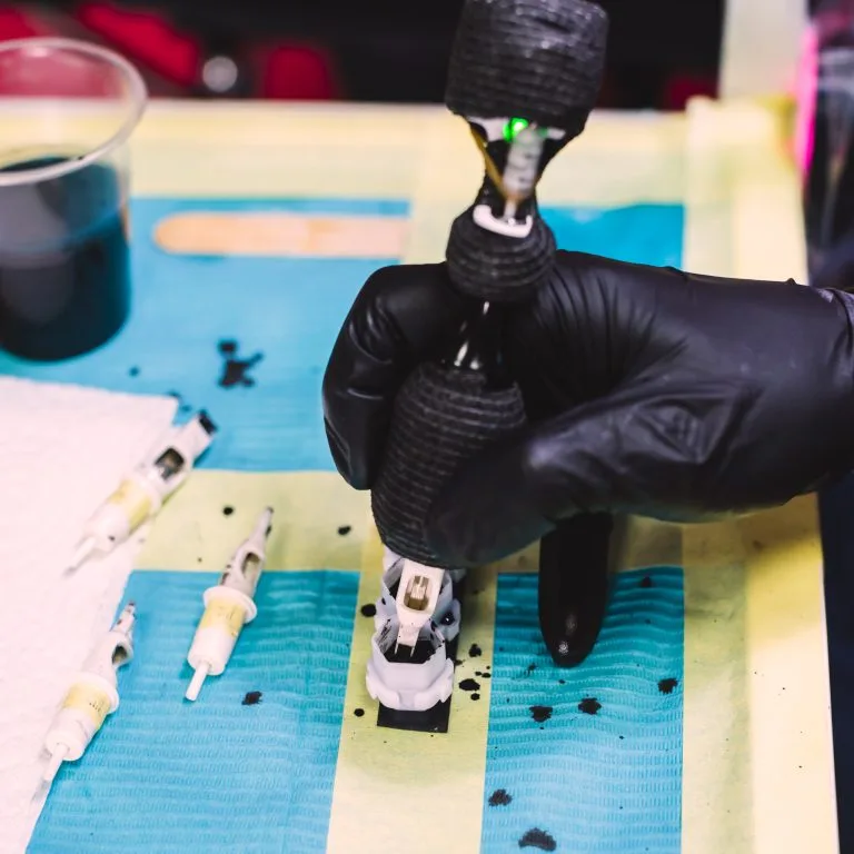 A gloved hand holds a tattoo machine near ink-stained needles and a cup of black ink on a blue protective surface, highlighting the tattoo placement process and what to expect during your session.