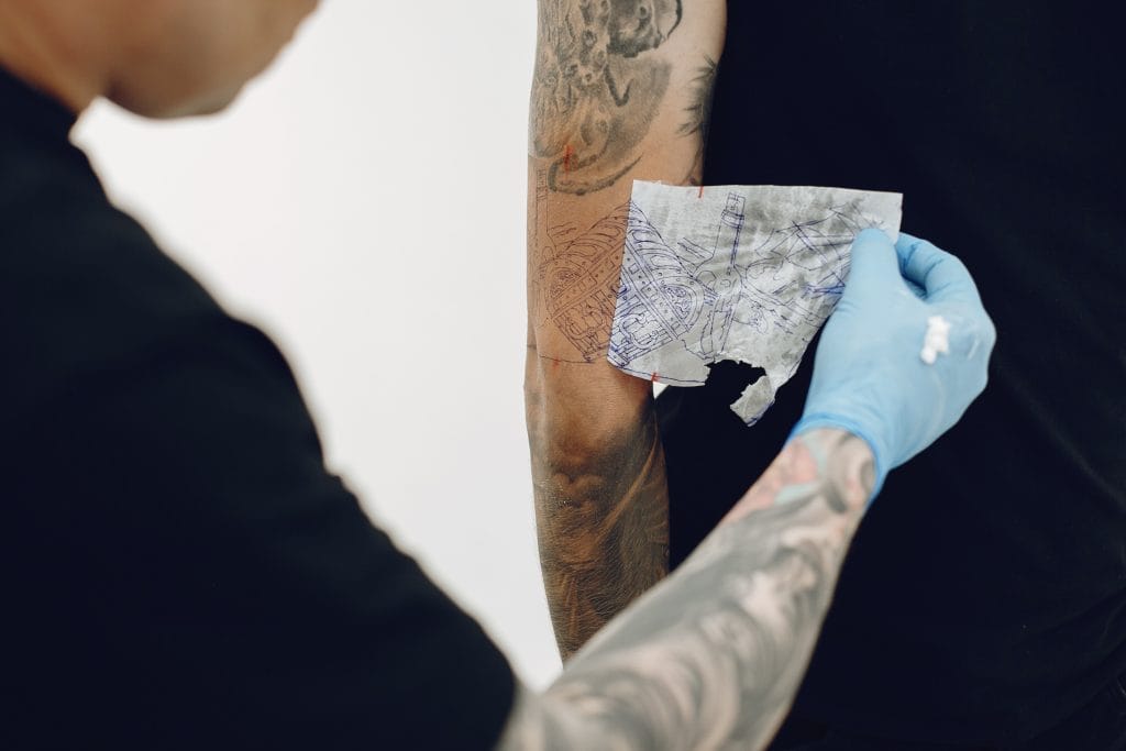 A tattoo artist in gloves applies a stencil design to a person's arm in preparation for a tattoo, emphasizing the importance of plant-based tattoo aftercare such as Tattoo Lotions or Tattoo Balms for optimal healing.