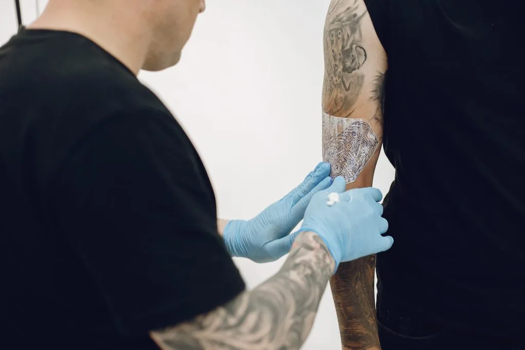 A tattoo artist wearing blue gloves applies a protective film to a freshly tattooed arm of a person dressed in black, highlighting the importance of plant-based tattoo aftercare for optimal healing.