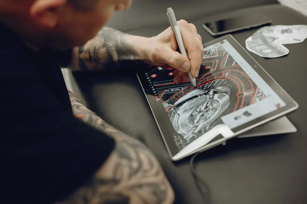 A person with tattooed arms uses a stylus to draw art sketches on a tablet displaying digital masterpieces; a phone and paper cutout are on the table nearby, capturing the creative spirit of Funhouse Artists.