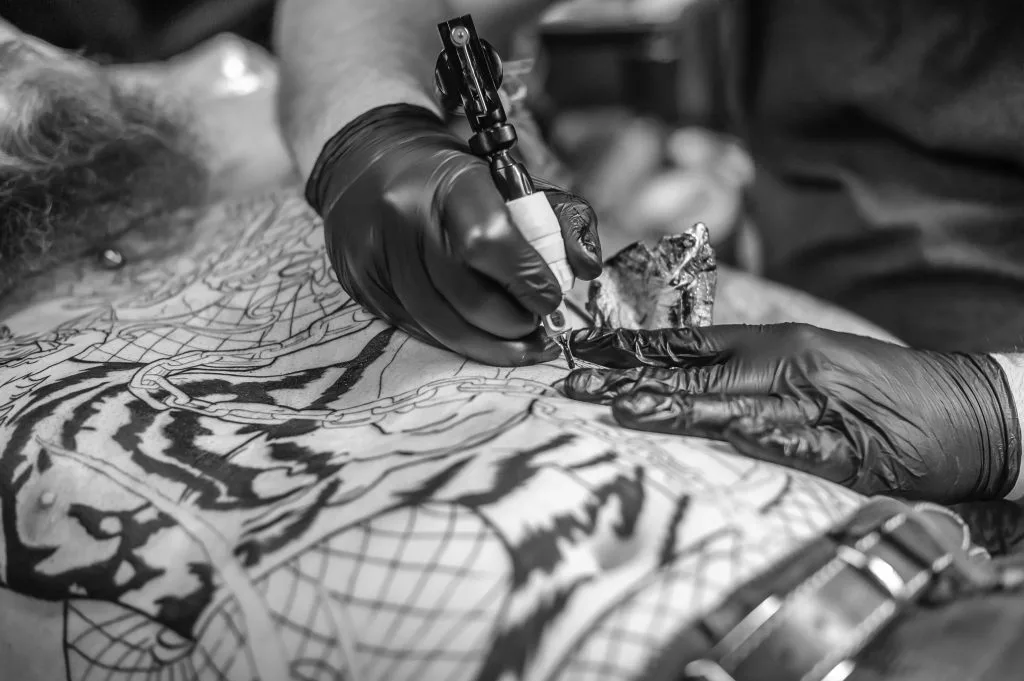 A tattoo artist from Funhouse Artists, wearing black gloves, uses a tattoo machine to ink a detailed masterpiece on a person's chest.