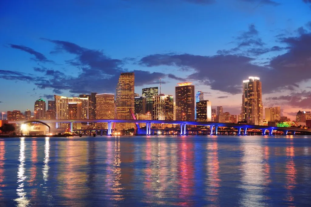 A city skyline at dusk with illuminated buildings, a lit-up bridge, and reflections of lights on calm water in the foreground—an inspiring scene reminiscent of San Diego tattoo culture and its iconic shops.