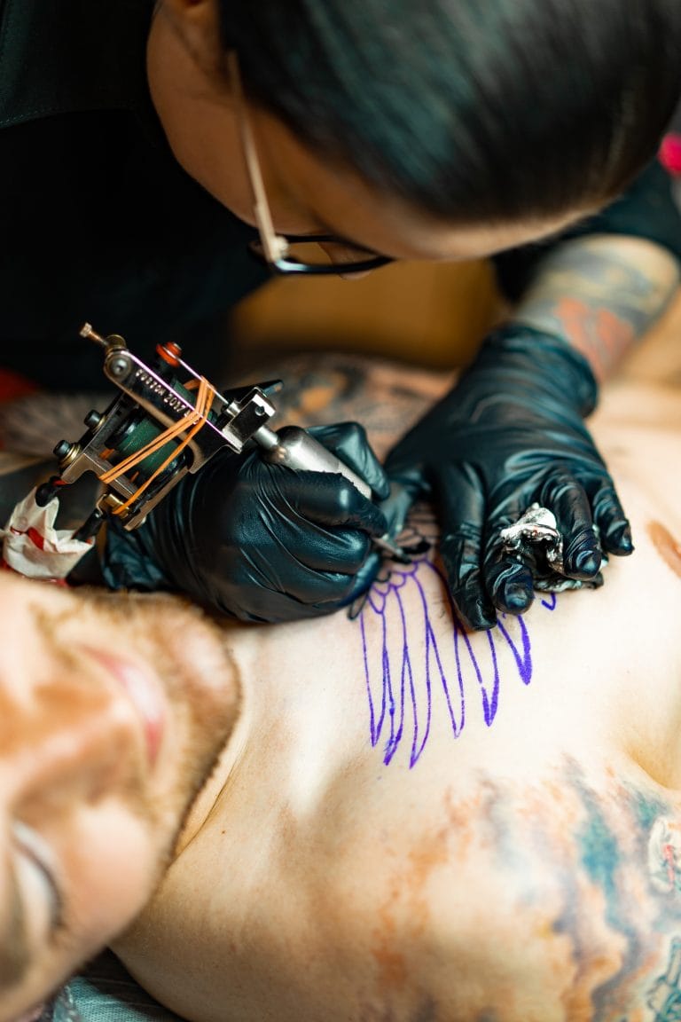 A tattoo artist wearing black gloves uses a tattoo machine to ink the outline of wings on a person's chest, focusing carefully on the Tattoo placement while considering the tattoo pain scale for this sensitive area.