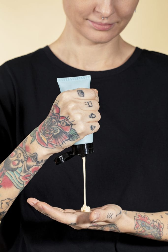 A person with tattoos on their arms dispenses tattoo lotion from a tube onto their hand, following a plant-based tattoo aftercare routine.