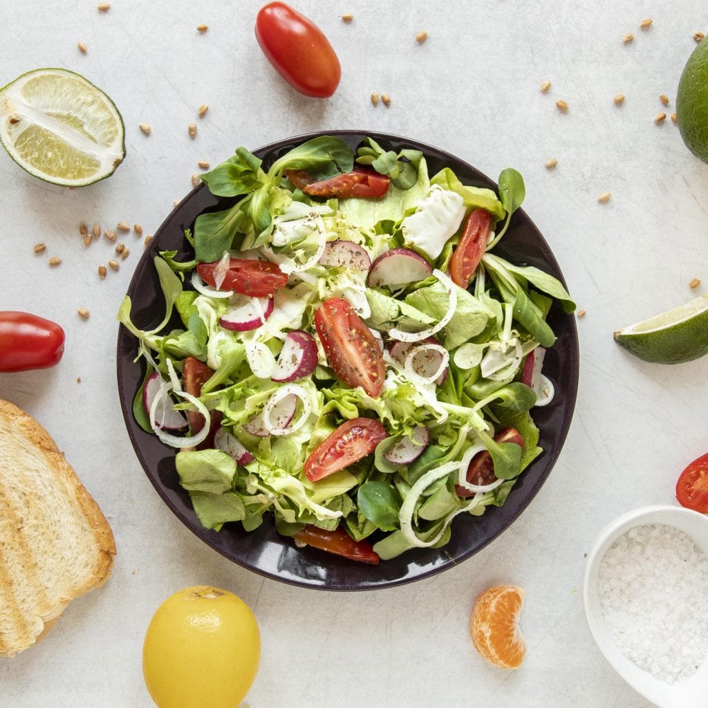 A bowl of fresh salad with tomatoes, radishes, and greens sits on a white surface, surrounded by bread, lemon, lime, salt, and other ingredients—perfect for nourishing your body before a full-day tattoo session.