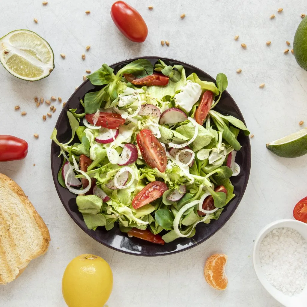 A bowl of fresh salad with tomatoes, radishes, and greens sits on a white surface, surrounded by bread, lemon, lime, salt, and other ingredients—perfect for nourishing your body before a full-day tattoo session.
