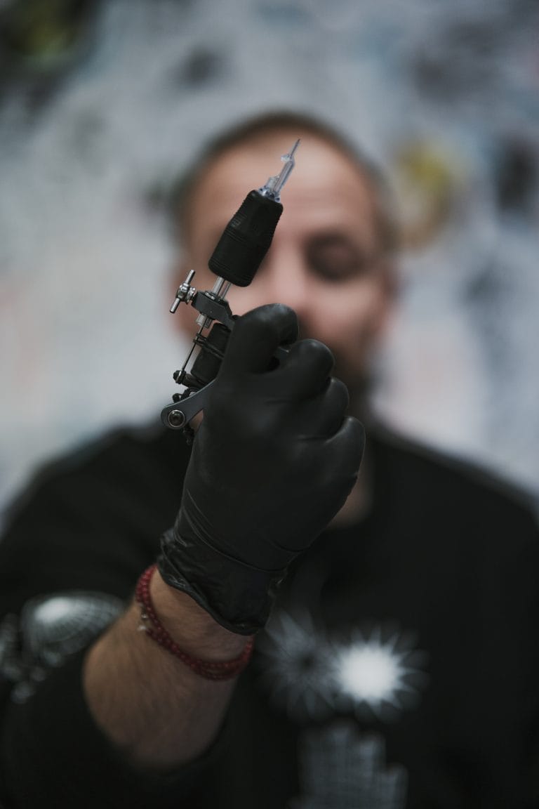 Person holding a tattoo machine with a needle, wearing black gloves and a black shirt.