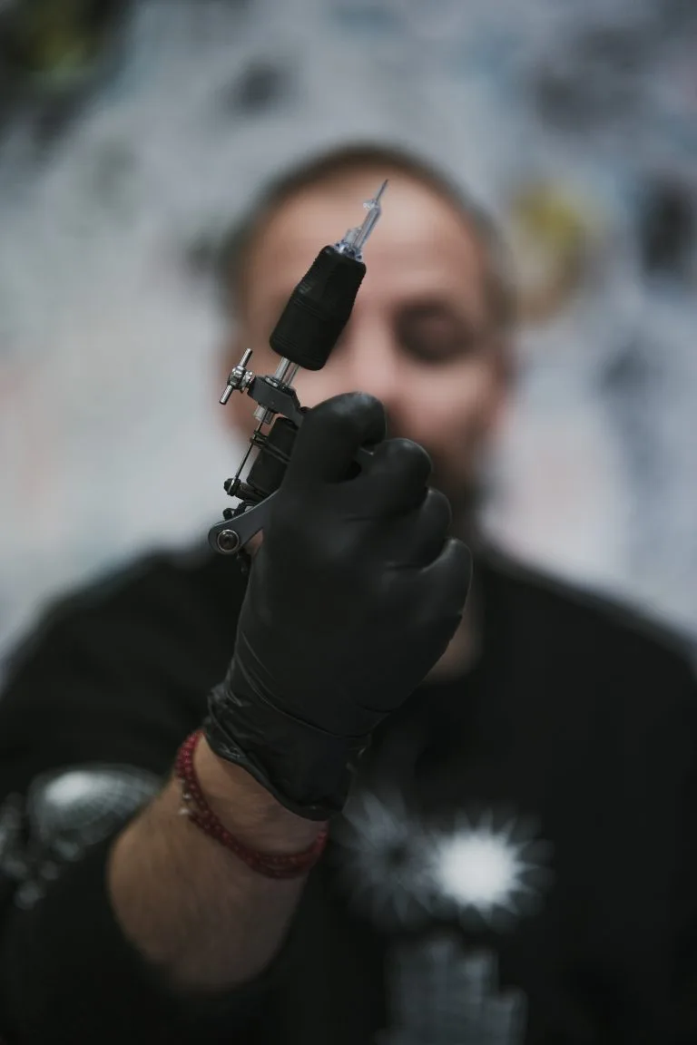Person holding a tattoo machine with a needle, wearing black gloves and a black shirt.