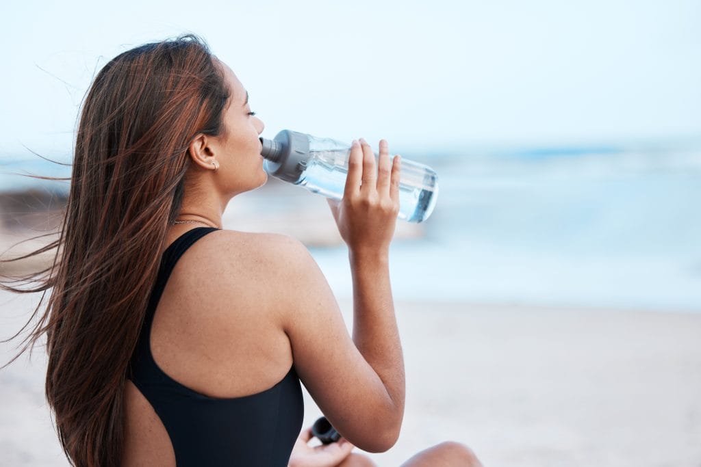 Woman in a black swimsuit drinks water from a bottle while sitting on the beach, practicing good tattoo preparation with hydration, as the ocean blurs in the background.