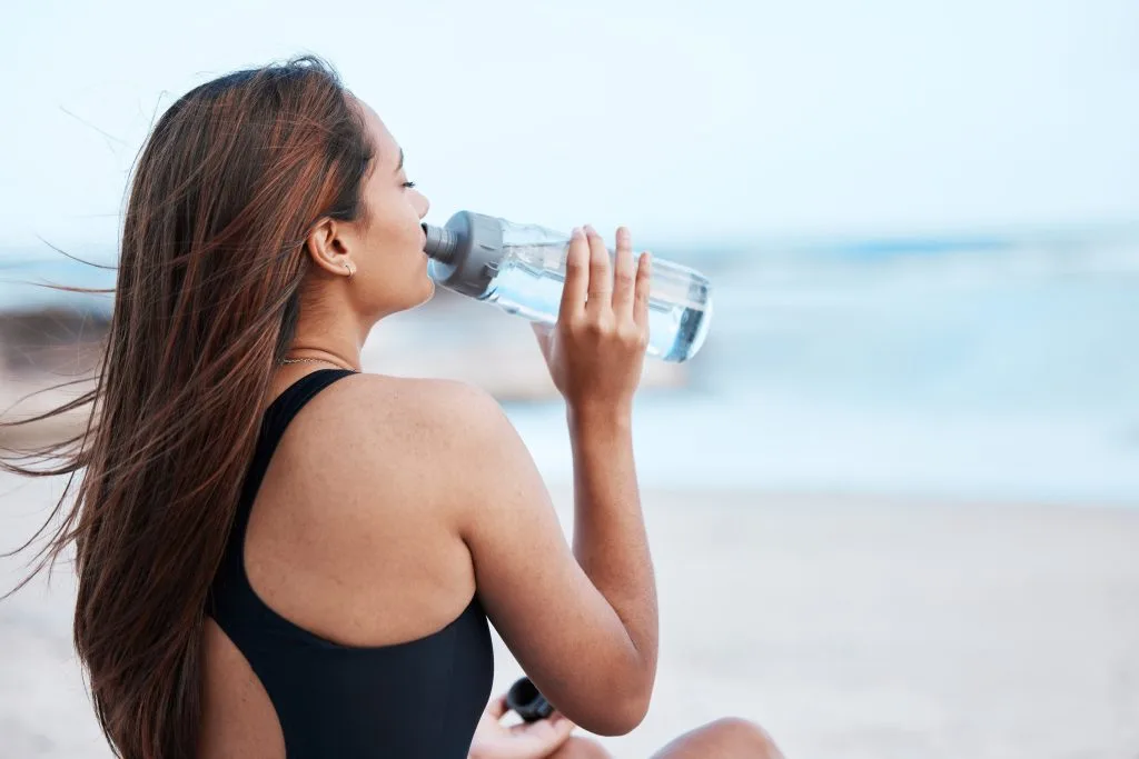 Woman in a black swimsuit drinks water from a bottle while sitting on the beach, practicing good tattoo preparation with hydration, as the ocean blurs in the background.