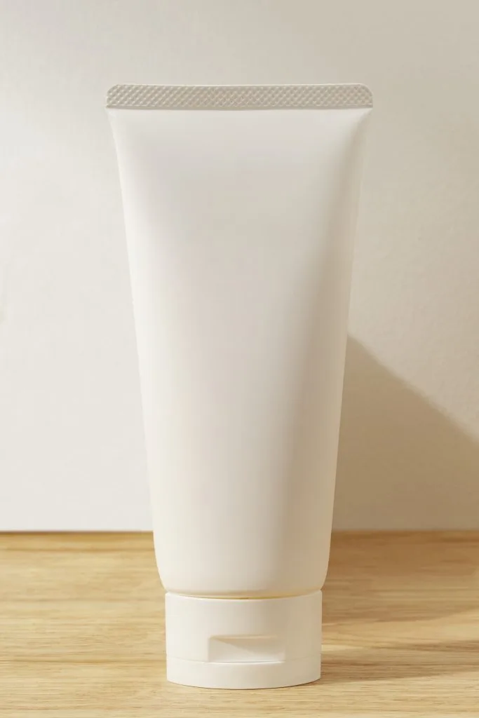 A plain, white, unbranded squeeze tube with a flip-top cap is standing upright on a wooden surface against a neutral background—perfect for storing tattoo lotions or plant-based tattoo aftercare products.