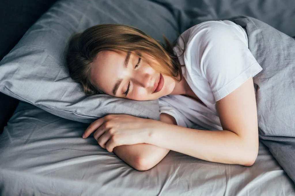 A woman with light brown hair is lying on her side in bed, sleeping, resting well as part of her tattoo preparation before a full-day tattoo session, with her head on a gray pillow and covered by a gray blanket.