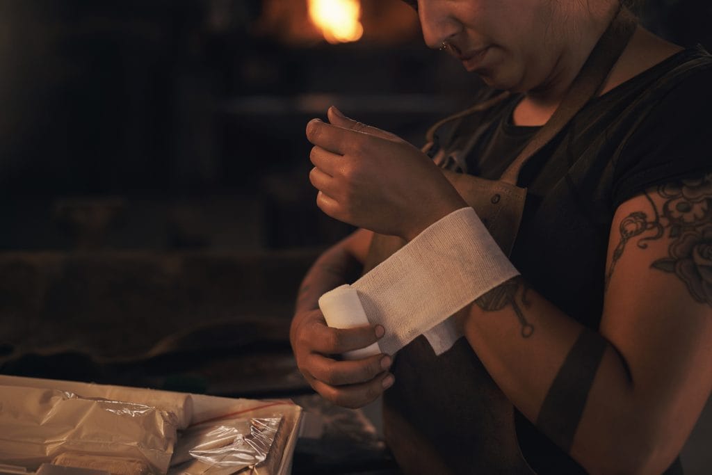 A person with tattoos wraps a bandage around their wrist, holding plant-based tattoo aftercare supplies on a dark workbench, with a blurred fire in the background.