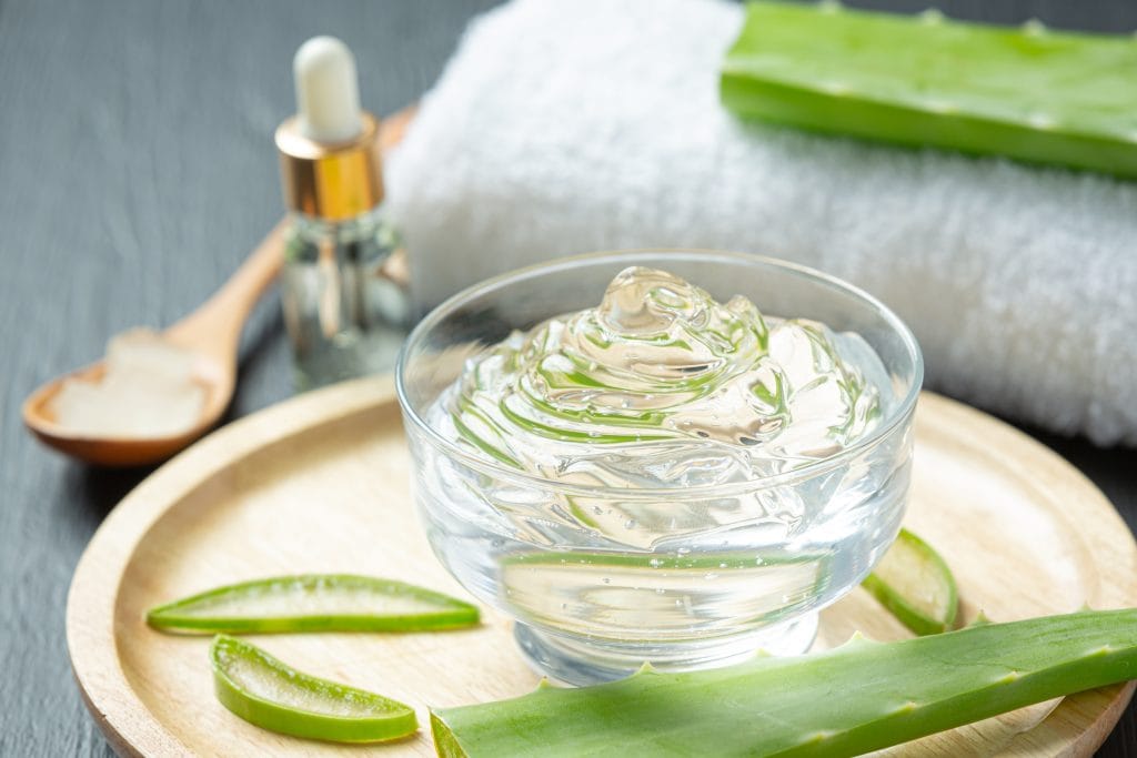 A glass bowl of aloe vera gel on a wooden tray, surrounded by fresh aloe vera leaves, a dropper bottle, a wooden spoon, and a rolled white towel—perfect for soothing tattoo reactions and sensitivities naturally.