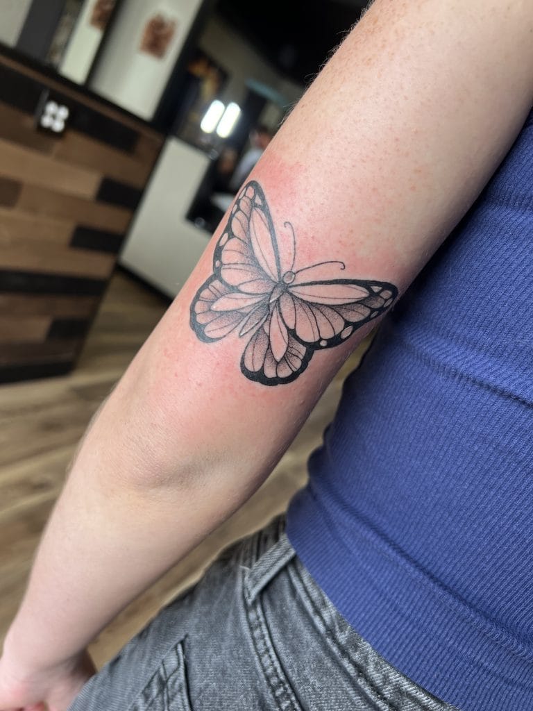 Black and gray butterfly tattoo on persons upper arm, with a pinkish background.