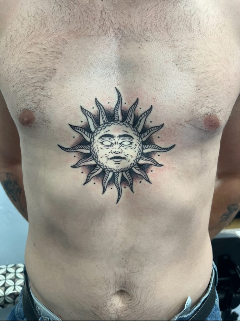 Tattoo of a sun with a human face and rays extending outward on the persons chest.
