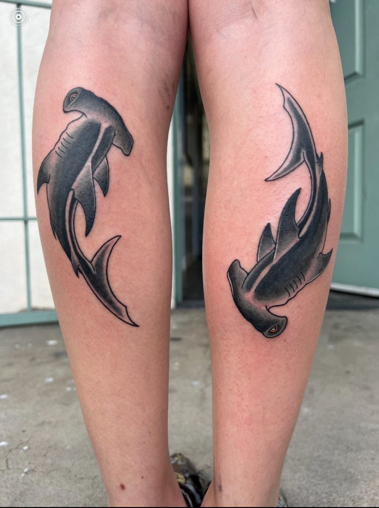 Two black and gray sharks tattooed on the backs of persons calves, facing opposite directions.