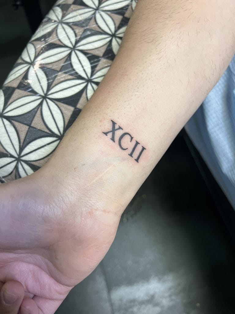 Roman numeral tattoo XCII on inner wrist, with a patterned surface nearby.