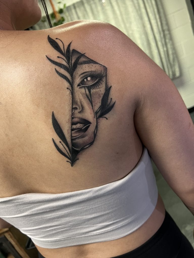Tattoo of a womans face with leaves and a tear, partially hidden by a vertical line.