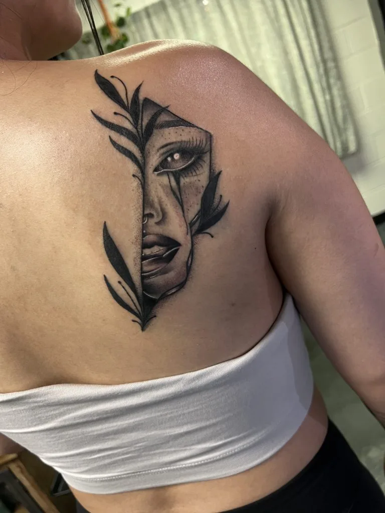 Tattoo of a womans face with leaves and a tear, partially hidden by a vertical line.