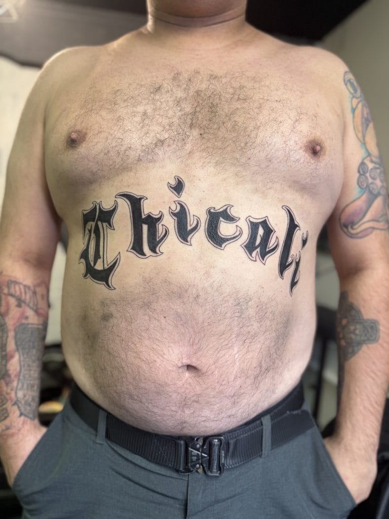 Man with chest tattoo reading Chivalry and colorful tattoos on arms, wearing gray pants.