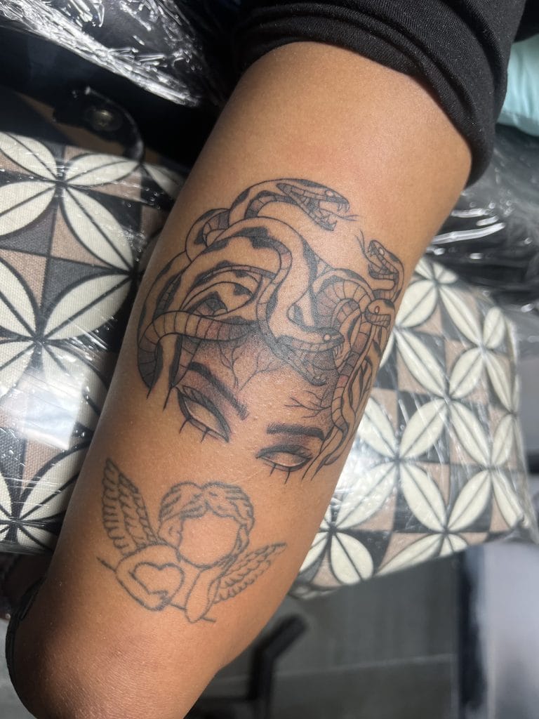 Tattoo of Medusa with snakes and a cherub with wings and a heart on forearm.