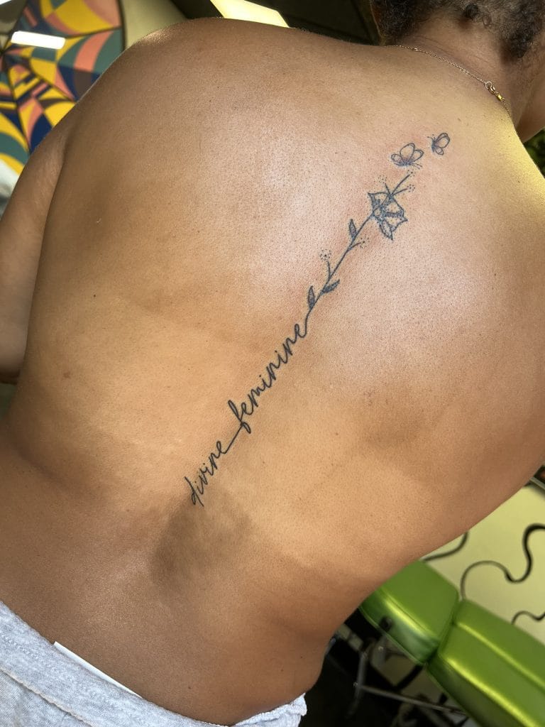 Vertical tattoo of a flower stem with butterflies and the words vive féminine on a persons back.