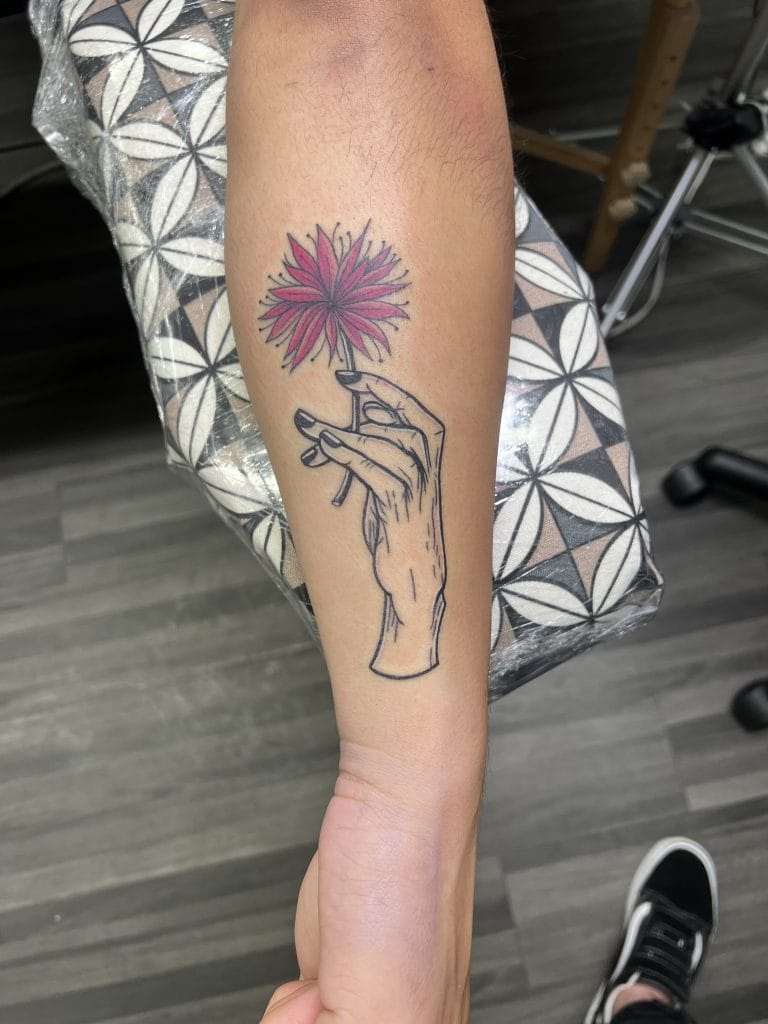 Hand holding a pink flower with black outline, tattooed on forearm.