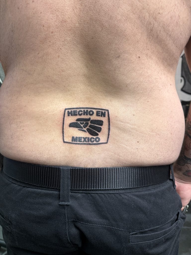 Tattoo on lower back with the text HECHO EN MEXICO and a fist symbol inside a rectangle.