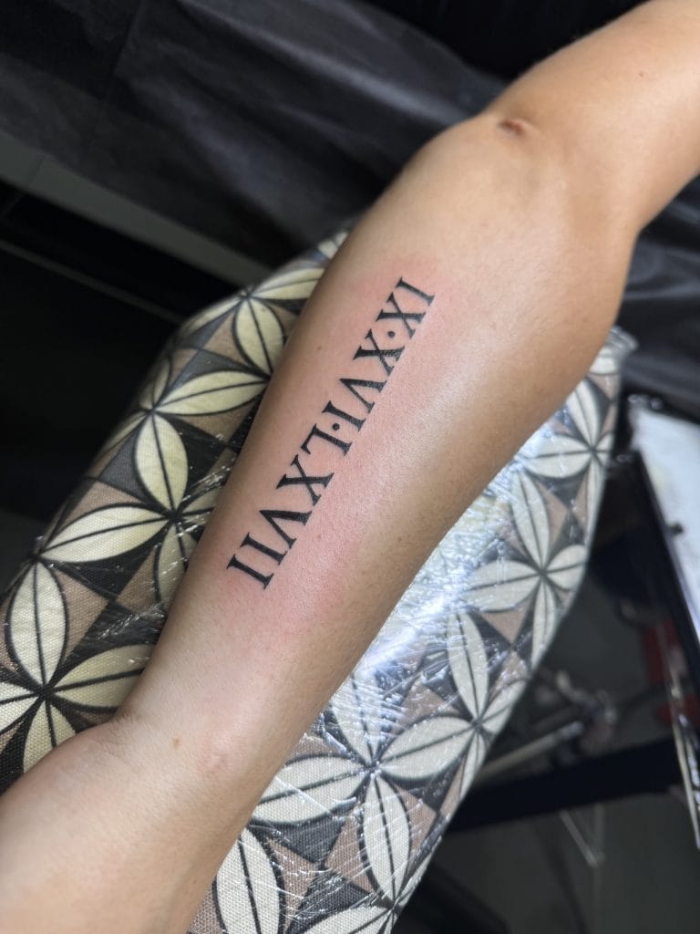 Roman numeral tattoo on forearm reading XIX:XXIX:XXI in black ink.