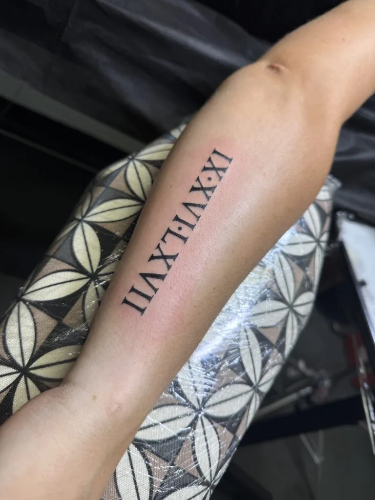 Roman numeral tattoo on forearm reading XIX:XXIX:XXI in black ink.