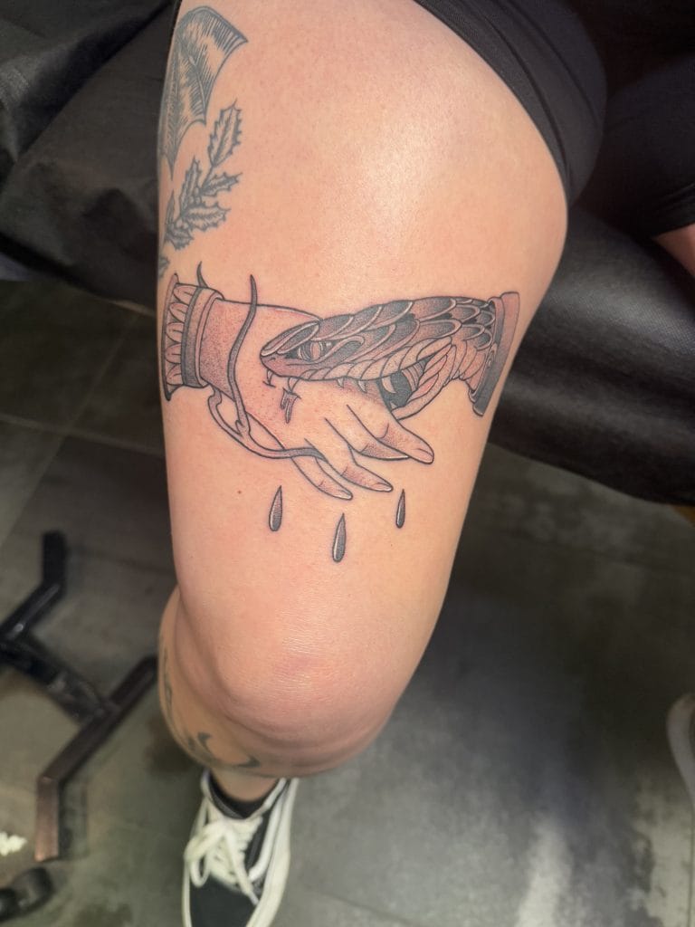 Hand holding a snake with drops falling from its mouth tattooed on a persons thigh.