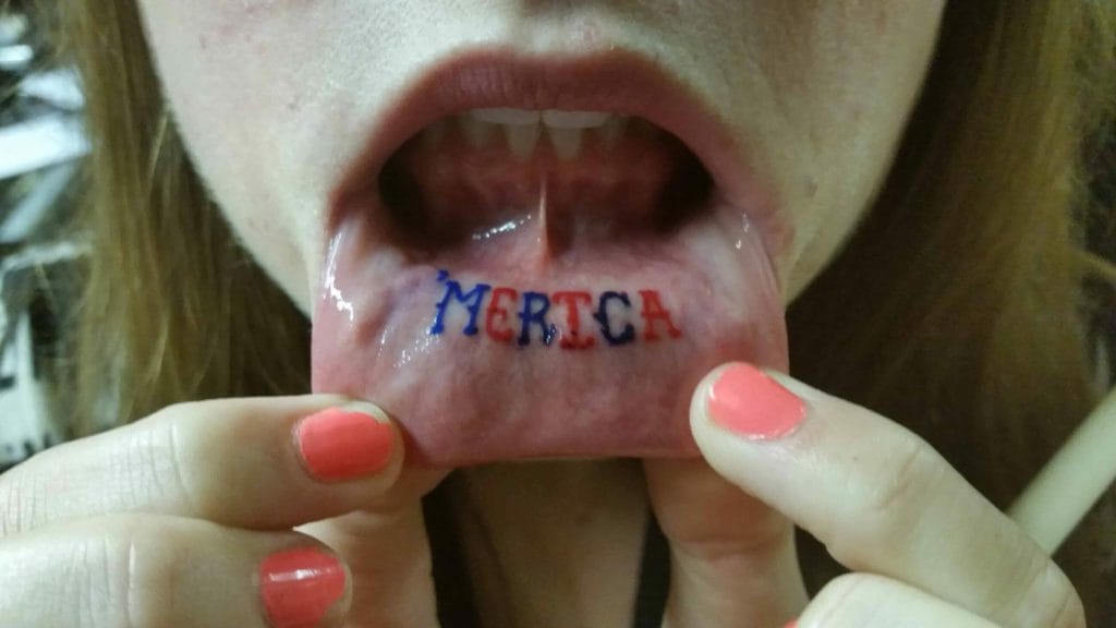 Person holding open mouth with MERTCA tattooed on tongue, showing teeth and lips.
